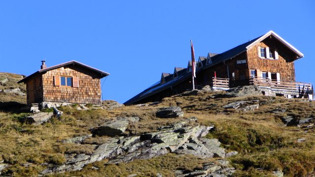 Badener Hütte des Alpenverein Baden auf 2.608 Metern: Schutzhaus mit Holzschindeln und kleines Nebengebäude auf felsigem Hang, blauer Himmel
