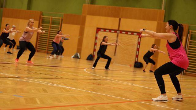 Fitnesskurs der ASKÖ Traiskirchen-Möllersdorf in einer Turnhalle, die Trainerin macht eine Kniebeuge mit gestreckten Armen vor, die Gruppe macht mit