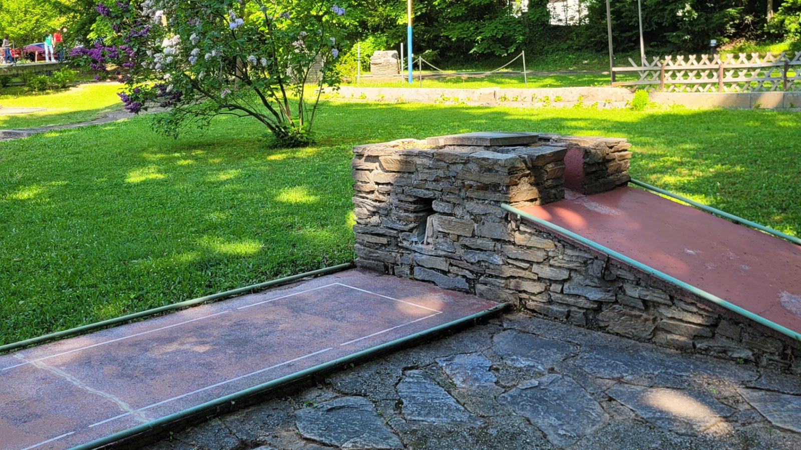 Minigolfbahnen des Badener AC am Kaiser-Franz-Ring mit Steinmauer und Rampe, dahinter Rasen, Flieder und hohe Laubbäume