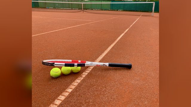 Tennisschläger und drei gelbe Tennisbälle liegen auf einem Sandplatz an der Linie, dahinter das Netz – Hintergrundbild des Badener Tennisclubs