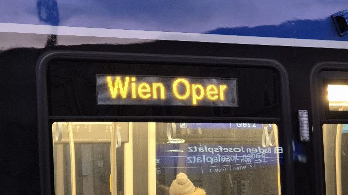 Front eines Zuges der Badner Bahn mit der gelben Zielanzeige „Wien Oper“; im Fenster darunter spiegelt sich die Bahnsteiganzeige „Baden Josefsplatz, Gleis 2“