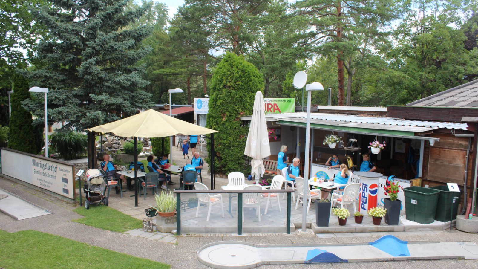 Minigolfanlage des BGSC Leobersdorf mit Bahn im Vordergrund, Kantinenterrasse mit Sonnenschirm und Gästen, dahinter hohe Bäume