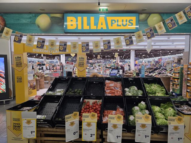 Eingang einer BILLA-PLUS-Filiale: gelber Schriftzug über der Tür, davor Obst- und Gemüsekisten mit Preisschildern „Immer clever Preis“