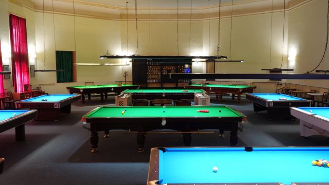 Clubsaal des Billard-Sportvereins Baden mit blauen Pooltischen und grünen Snookertischen, im Hintergrund Pokalschrank und rote Vorhänge