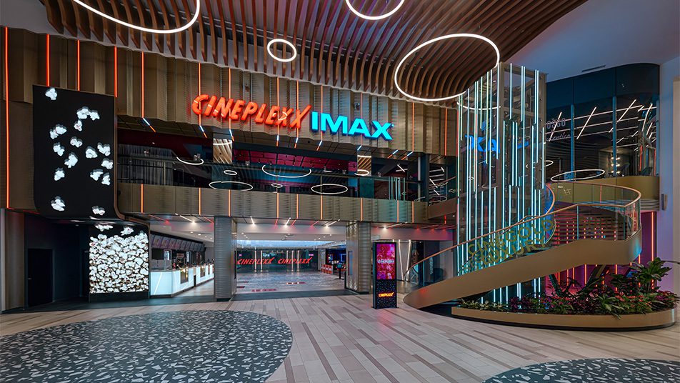 Foyer des Cineplexx Westfield SCS mit rotem Leuchtschriftzug „Cineplexx“ und blauem „IMAX“, geschwungener Treppe und runden Lichtringen an der Decke
