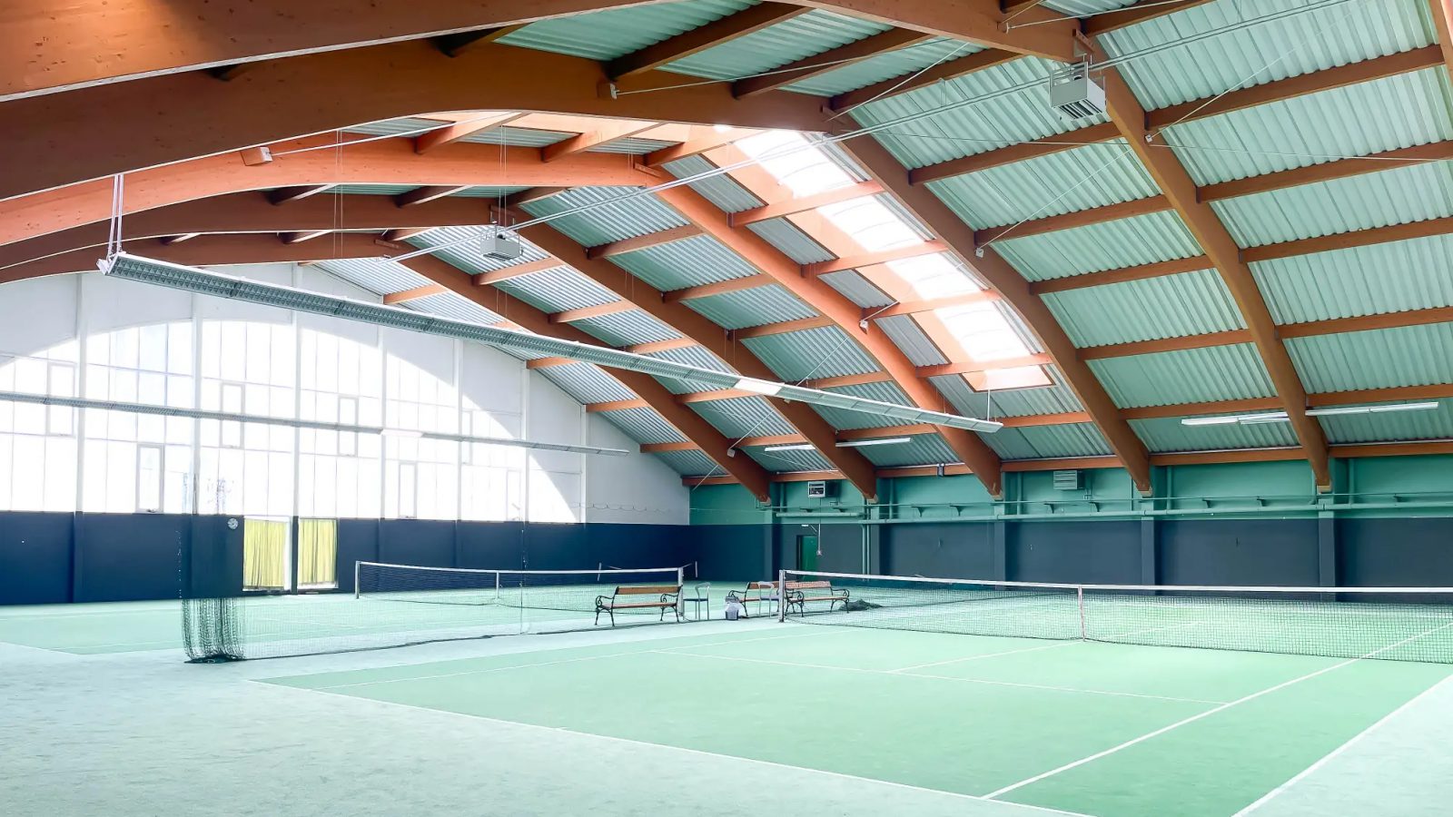 Tennishalle des Club Top & Fit in Bad Vöslau mit zwei grünen Teppichplätzen, Netzen, Bänken und einem Dach aus geschwungenen Holzbindern