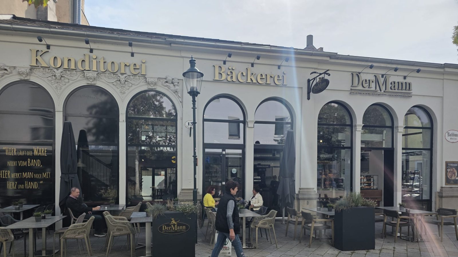 Die Filiale in der Rathausgasse 13 in Baden: helles Haus mit Rundbogenfenstern, darüber die goldenen Schriftzüge Konditorei, Bäckerei und Der Mann, davor der Schanigarten mit Tischen und Stühlen
