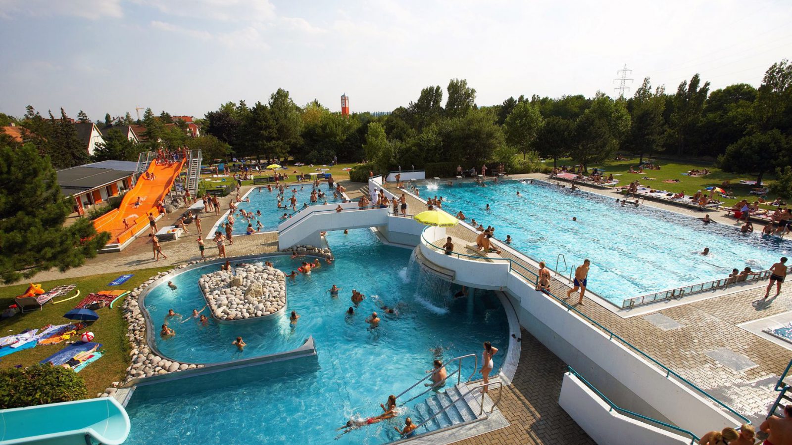 Erlebnisbad Leobersdorf von oben: Erlebnisbecken mit Wasserfall, Sportbecken, orange Breitrutsche und Liegewiese voller Badegäste