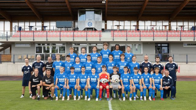 Mannschaft des FCM Traiskirchen in blauen Trikots mit Trainern und Betreuern beim Teamfoto vor der Tribüne der ARBÖ-Arena