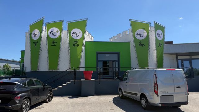 Eingang von fit+ Leobersdorf: weißes Gebäude mit grünem Eingangsbereich und fünf grünen fit+-Fahnen, davor der Parkplatz