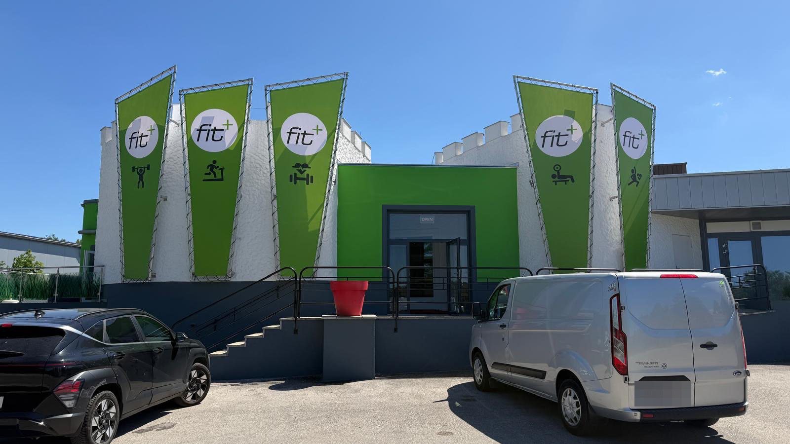 Eingang von fit+ Leobersdorf: weißes Gebäude mit grünem Eingangsbereich und fünf grünen fit+-Fahnen, davor der Parkplatz