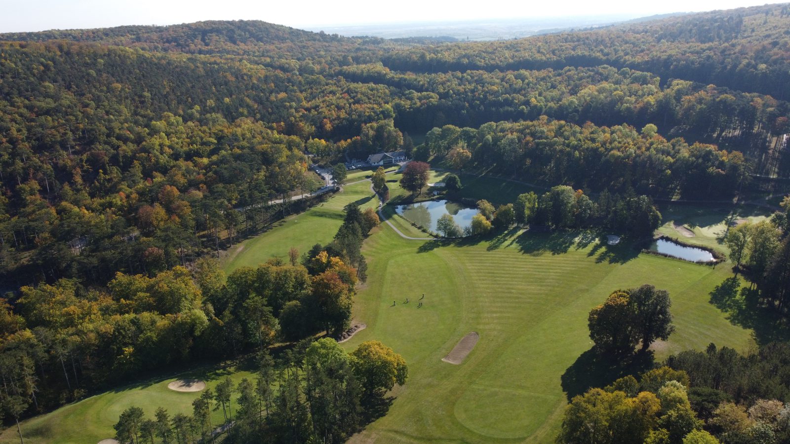 Golfplatz Enzesfeld aus der Luft im Herbst: Fairways und Teiche zwischen bewaldeten Hügeln, hinten das Clubhaus mit Parkplatz
