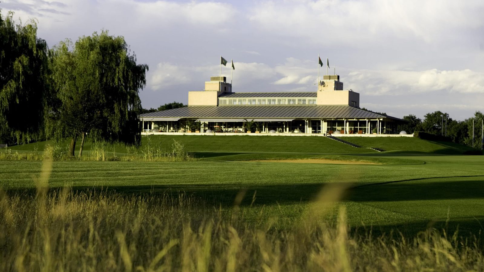 Golfplatz Schloss Ebreichsdorf: Blick über das Fairway des 18. Lochs mit Bunker auf das Clubhaus mit Glasfront und Fahnen