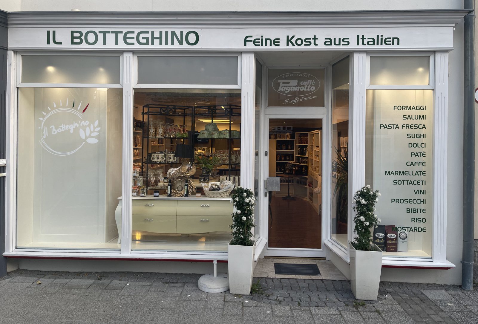Schaufenster von Il botteghino mit dem Schriftzug „Feine Kost aus Italien“, offener Eingangstür und einer Liste der Waren wie Formaggi, Salumi, Pasta fresca und Vini