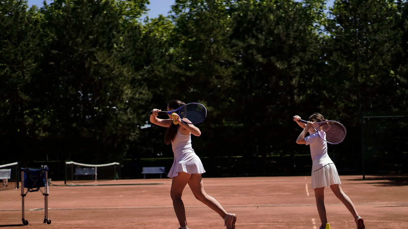 Zwei Tennisspielerinnen in weißer Kleidung üben auf einem Sandplatz die Rückhand, daneben Ballkorb und gelbe Hütchen, im Hintergrund Bäume