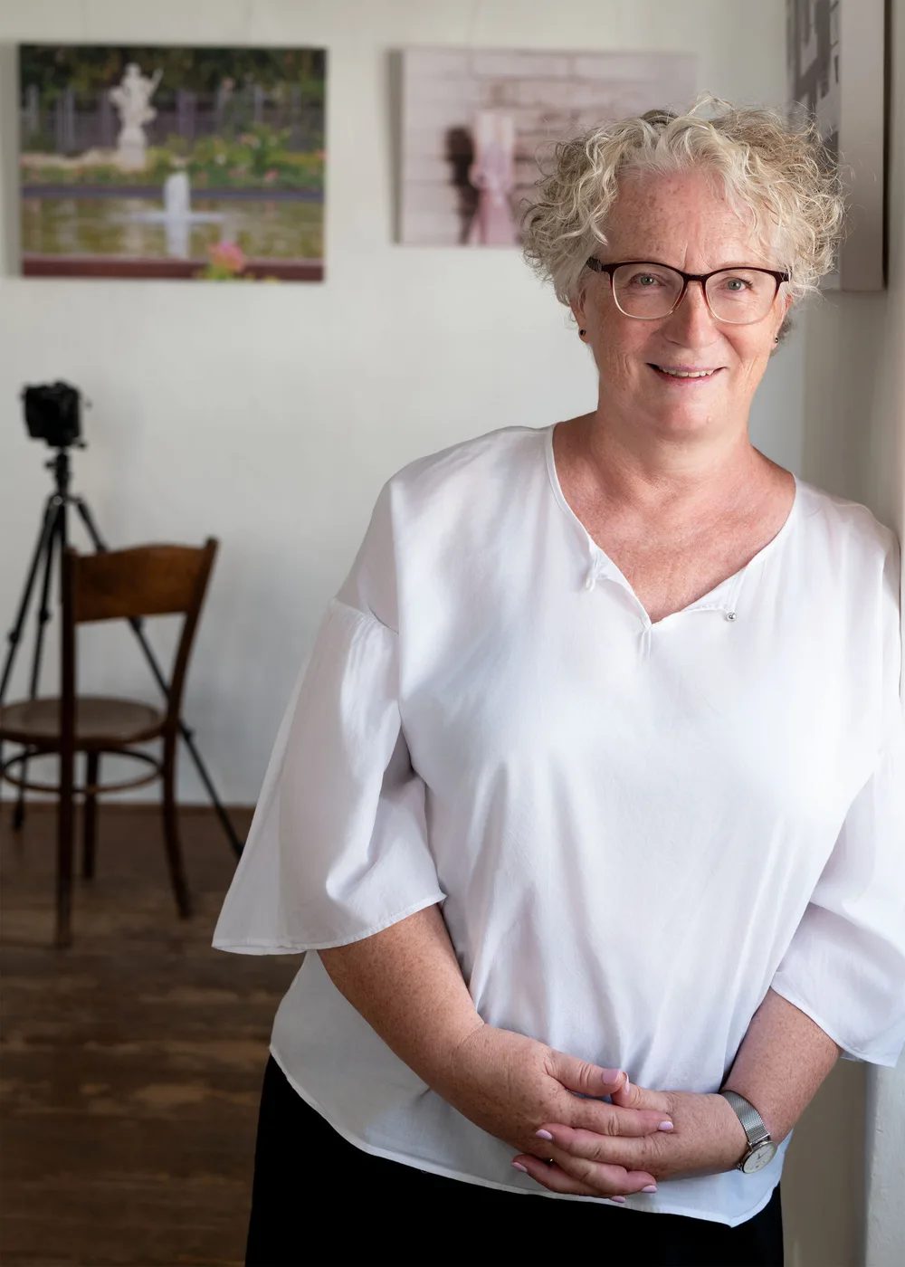 Michaela Vondruska, Fotografin aus Gumpoldskirchen, in weißer Bluse an eine Wand gelehnt, im Hintergrund eine Kamera auf dem Stativ und zwei ihrer Fotografien