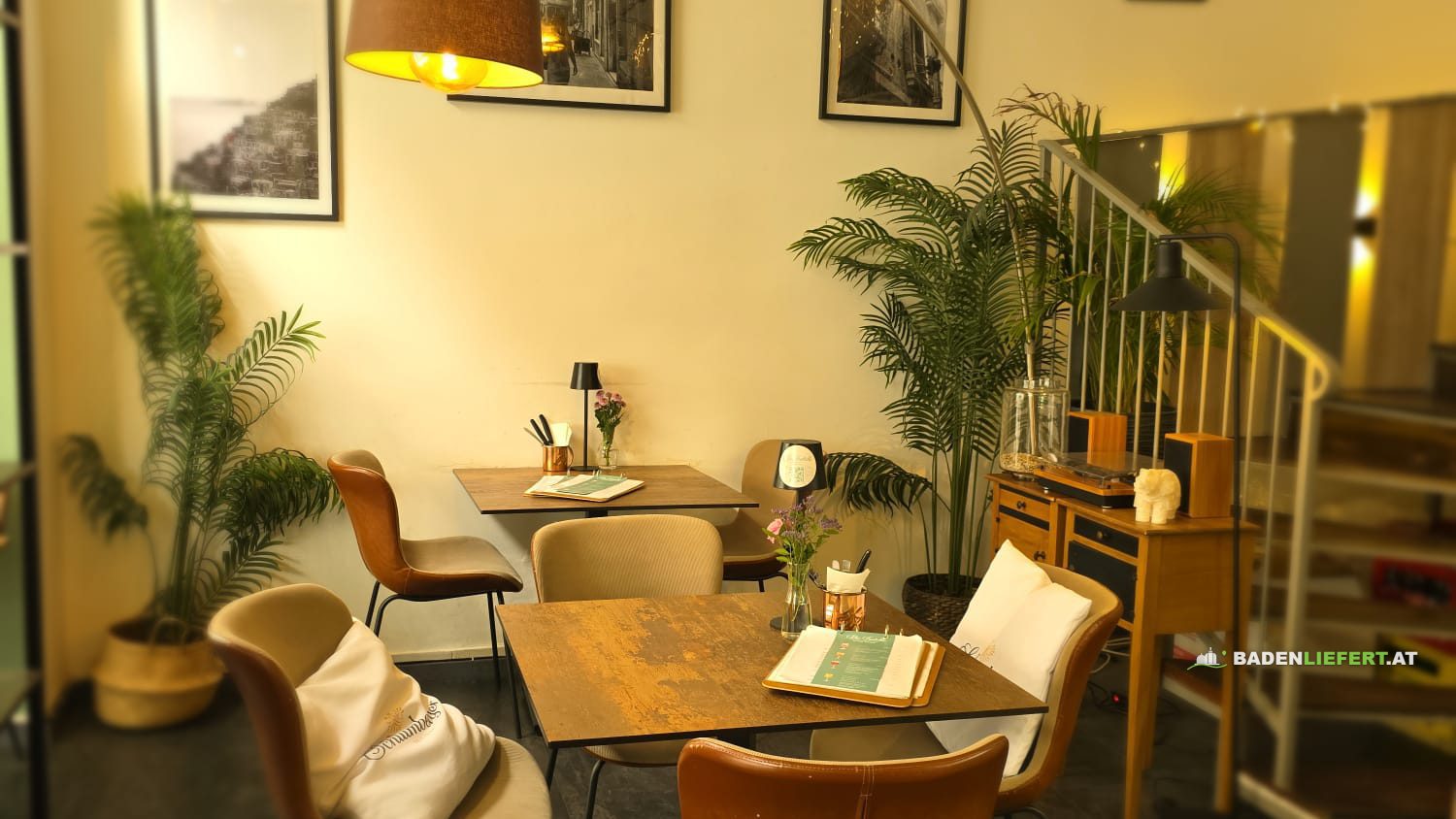 Gastraum von Mio Fratello: zwei Tische mit Lederstühlen, Bogenlampe, Palmen und schwarz-weiße Fotos an der Wand, rechts eine Treppe zur Galerie