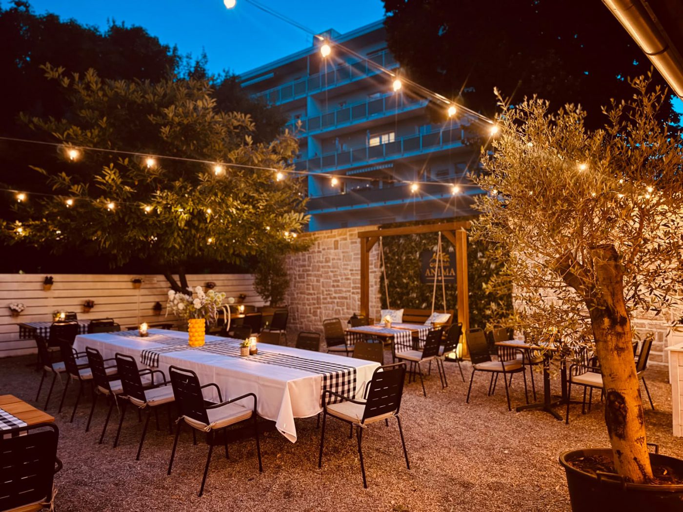 Gastgarten der Osteria Anima in der Dämmerung: eine lange gedeckte Tafel mit weißer Decke, darüber Lichterketten, rechts ein Olivenbaum im Topf