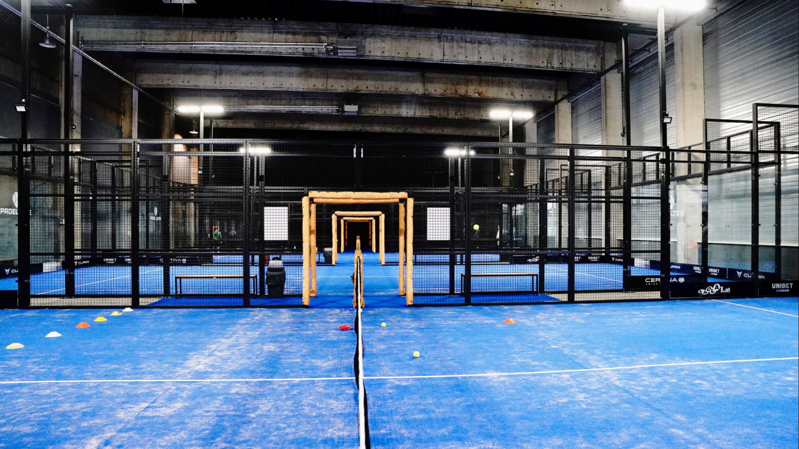 Blaue Indoor-Padelcourts der PADELZONE Traiskirchen mit Netz, Trainingshütchen und Bällen, dahinter weitere Courts unter einer Betondecke