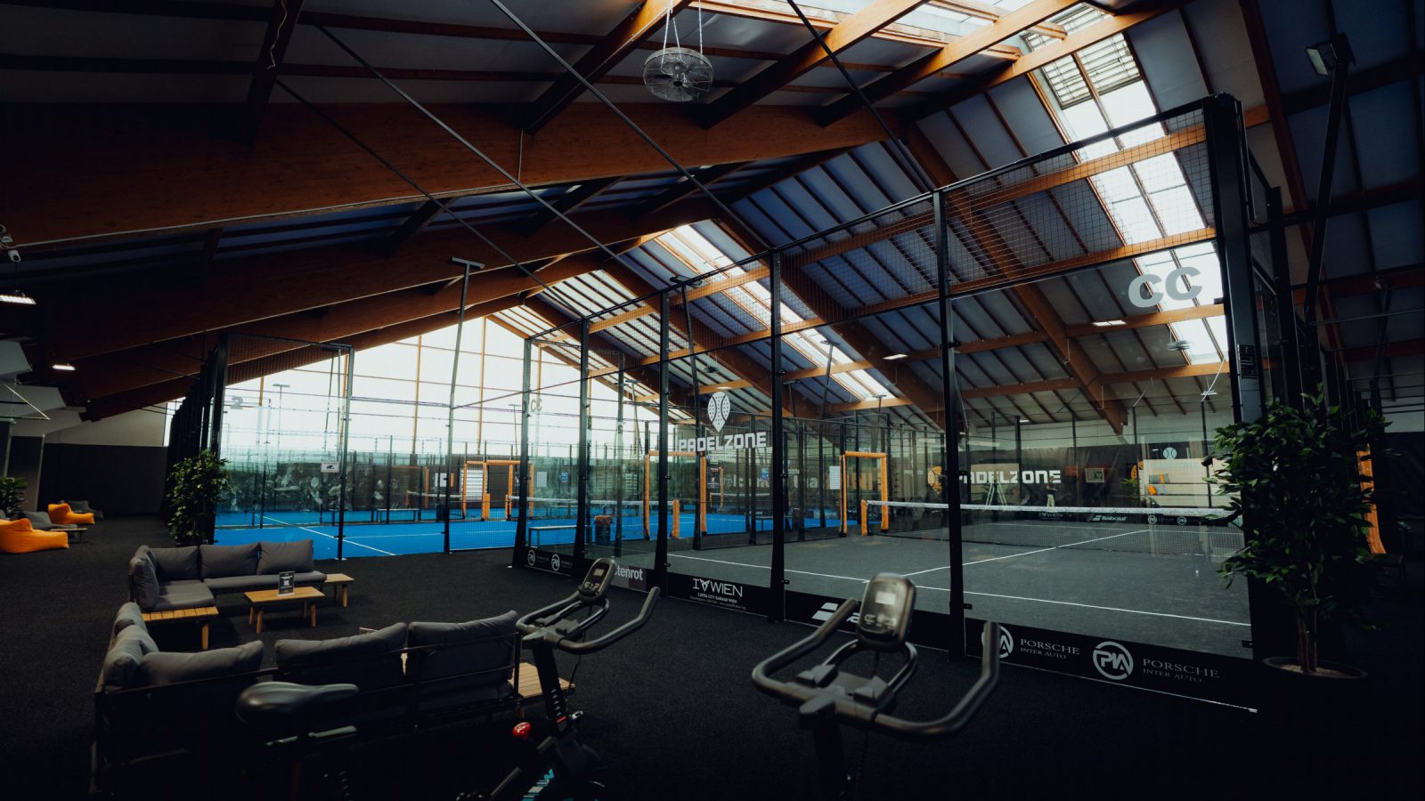Padelhalle der PADELZONE Vösendorf mit Holzdach und Oberlichtern, verglasten Courts und einer Lounge mit grauen Sofas im Vordergrund