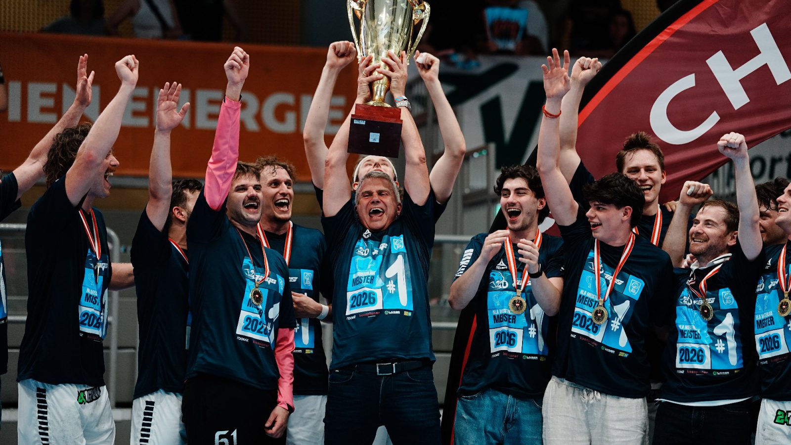 Spieler von roomz JAGS Vöslau feiern mit Medaillen und Meister-Shirts den HLA-Meistertitel 2026, in der Mitte wird der Pokal hochgehalten