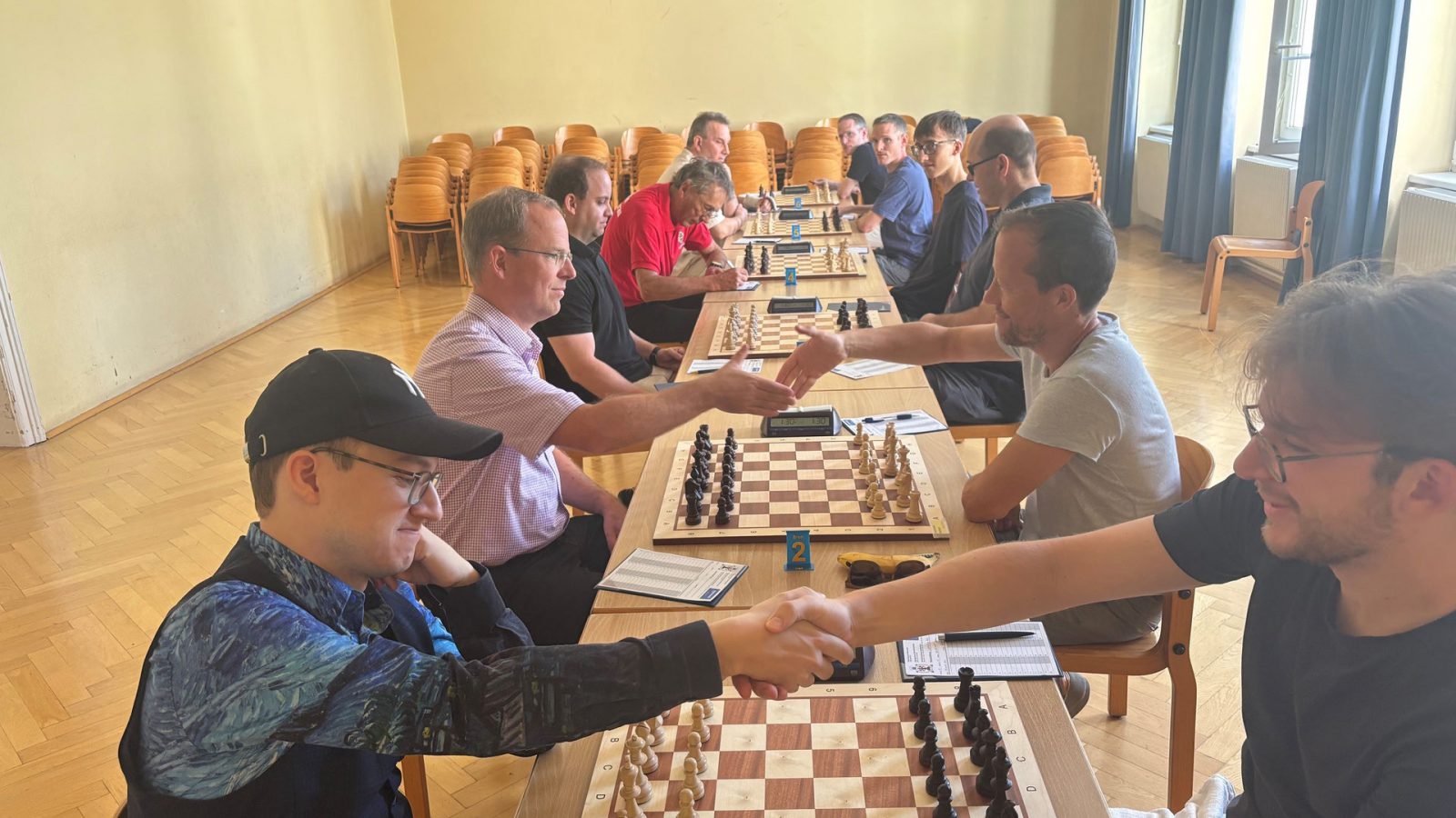 Schachspieler reichen sich an einer langen Tischreihe mit Schachbrettern und Uhren die Hand, Beginn eines Mannschaftskampfs des Schachklubs Baden