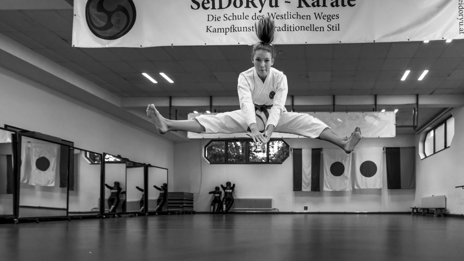 Karateka im weißen Karateanzug springt mit gegrätschten Beinen durch den Trainingsraum, darüber das Banner des Vereins SeiDoRyu