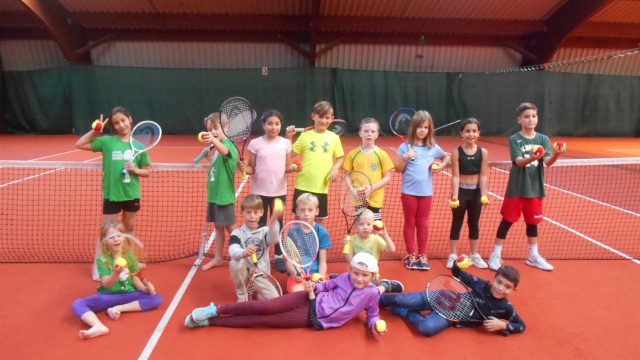 Kindergruppe mit Tennisschlägern und Bällen posiert am Netz eines roten Hallenplatzes in der Tennishalle des Sportcenters Baden