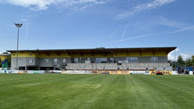 Sportzentrum Ebreichsdorf: Fußballplatz vor dem gelb-grauen Hauptgebäude mit Tribüne, links der Kletterturm, blauer Himmel