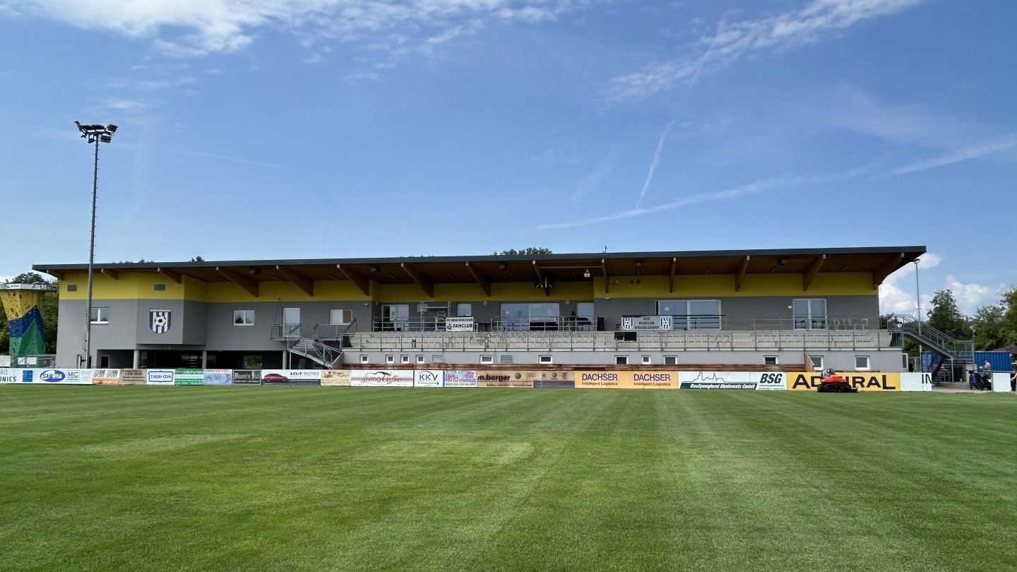Sportzentrum Ebreichsdorf: Fußballplatz vor dem gelb-grauen Hauptgebäude mit Tribüne, links der Kletterturm, blauer Himmel