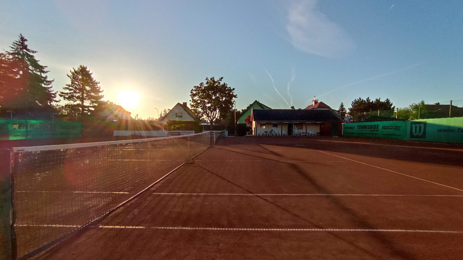 Sandplatz des TC ATUS Leobersdorf im tiefen Sonnenlicht, im Hintergrund das Klubhaus mit Terrassenstühlen und Bäume
