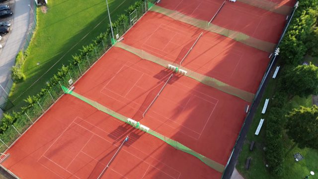 Luftbild der neuen Tennisplätze des TC Traiskirchen mit rotem Keramiksand-Belag, Netzen und Bänken, daneben Wiese, Hecken und Bäume