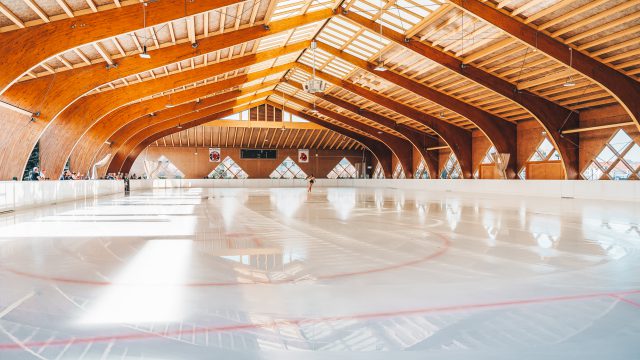 Kunsteisbahn im Sportzentrum Traiskirchen mit geschwungenem Holzdach und spiegelnder Eisfläche, im Hintergrund trainiert eine Eiskunstläuferin