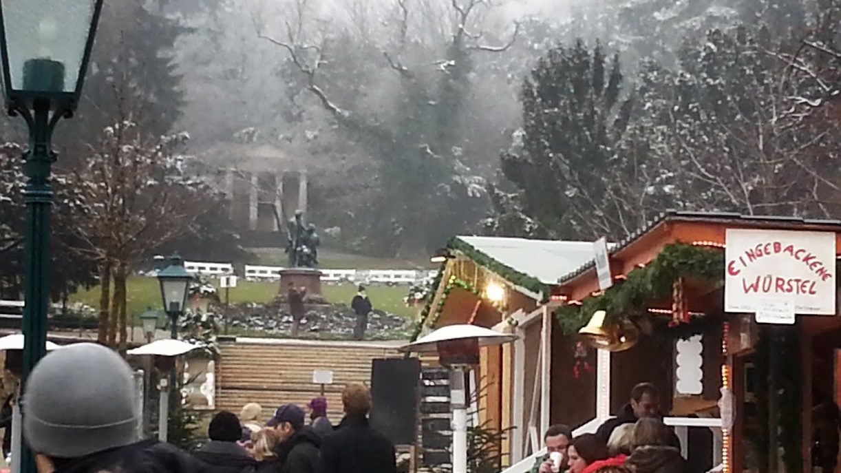 Adventmarkt im winterlichen Park: geschmückte Holzhütten mit Lichterketten und dem Schild „Eingebackene Würstel“, davor Besucher mit Punschbechern, im Nebel ein Denkmal und raureifbedeckte Bäume