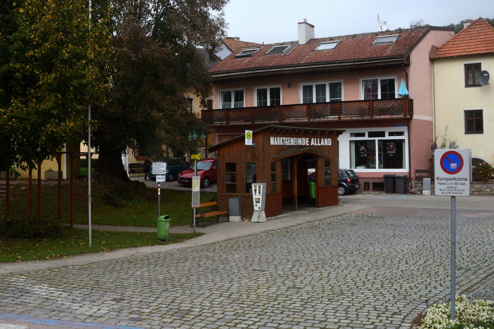 Gepflasterter Platz mit einem Holzhäuschen mit der Aufschrift Marktgemeinde Alland, dahinter ein Wohnhaus mit Balkon und Bäume im Herbstlaub