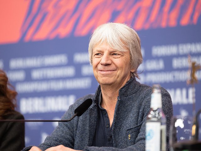 Ein Mann mit weißem Haar und grauer Strickjacke sitzt lächelnd an einem Tisch mit Mikrofon vor einer blauen Berlinale-Wand