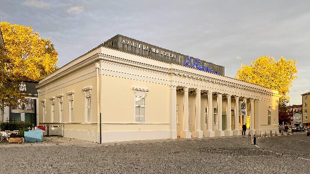 Klassizistischer, hellgelber Bau mit Säulenreihe, auf dem Dach der Schriftzug „Arnulf Rainer Museum“ und eine blaue Leuchtschrift, links und rechts Bäume mit gelbem Herbstlaub