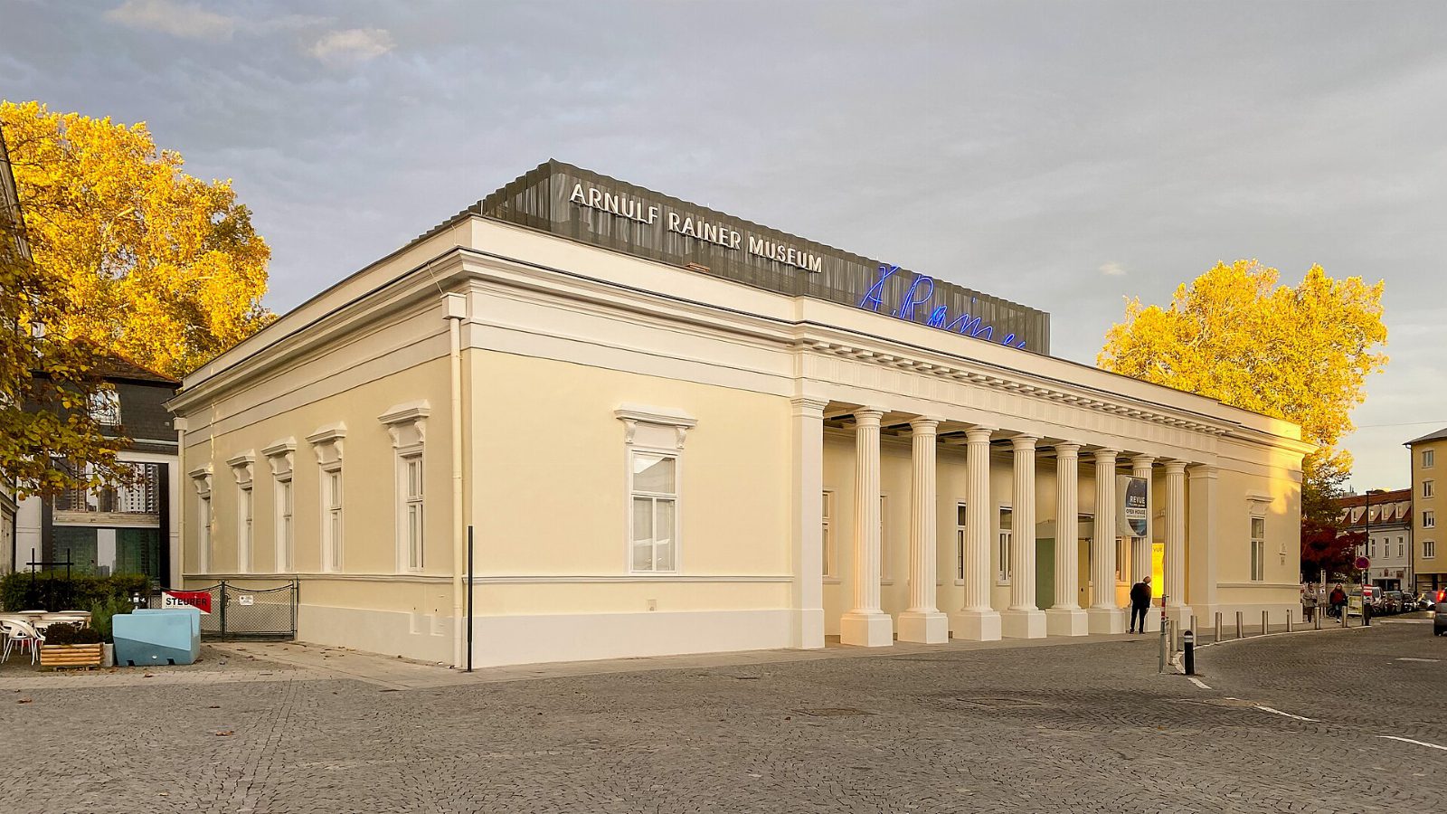 Klassizistischer, hellgelber Bau mit Säulenreihe, auf dem Dach der Schriftzug „Arnulf Rainer Museum“ und eine blaue Leuchtschrift, links und rechts Bäume mit gelbem Herbstlaub