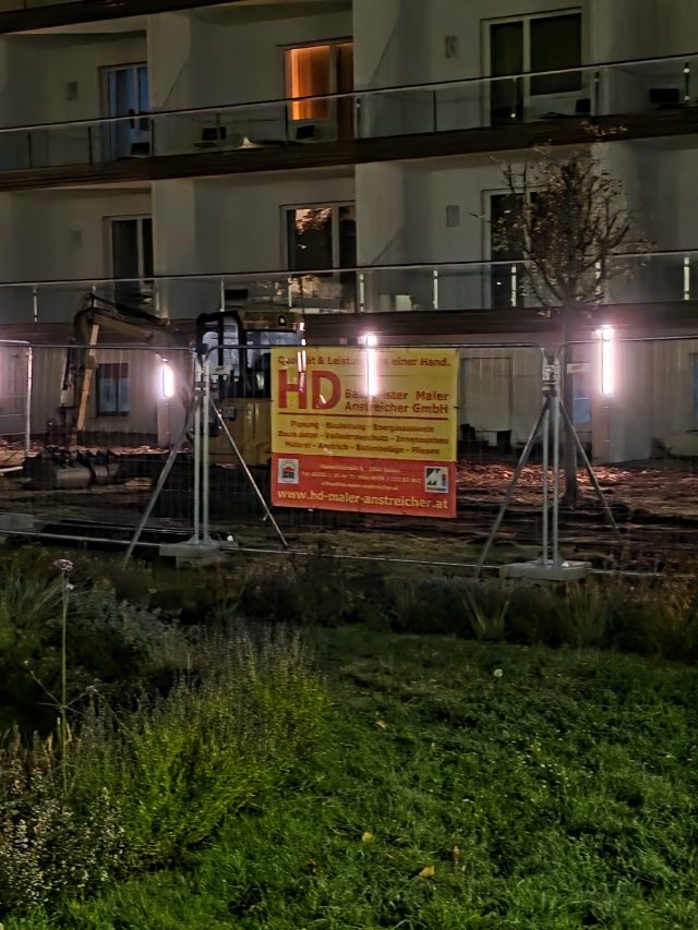 Baustelle vor dem At the Park Hotel in Baden bei Nacht: ein Bagger hinter dem Bauzaun mit gelb-roter Firmentafel, dahinter die Hotelfassade mit Glasbalkonen und Wandleuchten