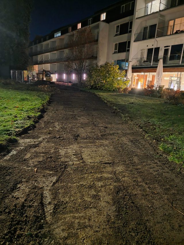 Abgetragener Rasenstreifen aus brauner Erde, der durch die Wiese zum Bauzaun und zum Bagger vor dem At the Park Hotel führt, rechts die beleuchtete Gartenfront des Hotels