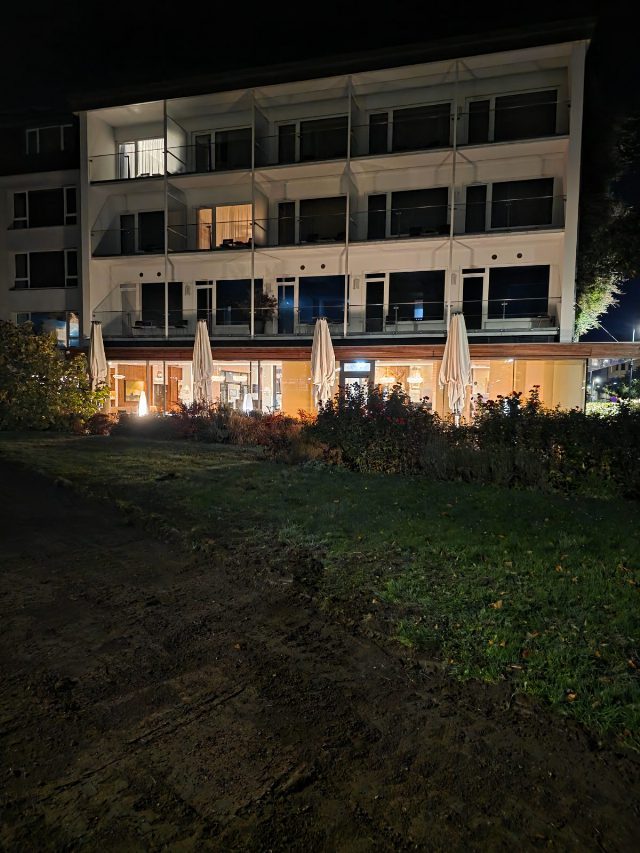 Gartenseite des At the Park Hotel in Baden am Abend: weiße Fassade mit Glasbalkonen, im Erdgeschoß das hell beleuchtete Restaurant hinter einer Glasfront, davor zusammengeklappte Sonnenschirme, Rosenbeete und Rasen