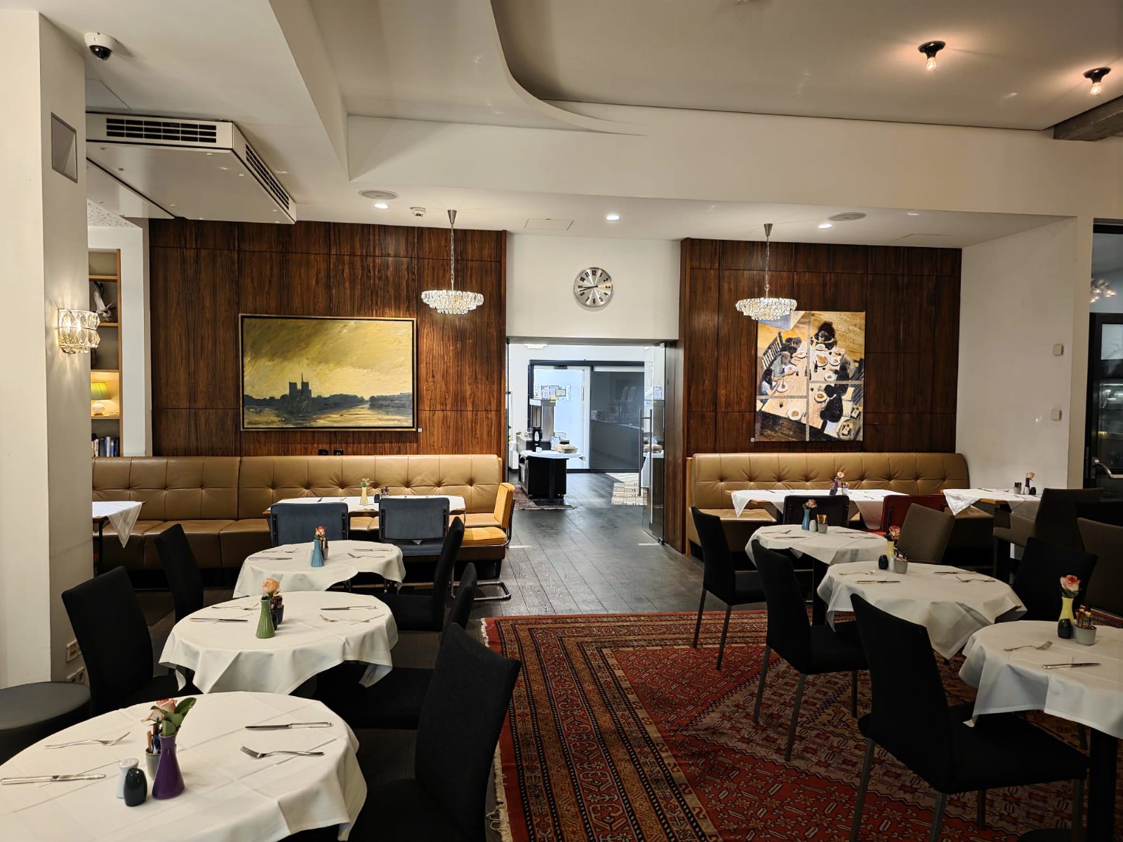 Speisesaal des At the Park Hotel in Baden mit runden, weiß gedeckten Tischen, Lederbänken an holzvertäfelten Wänden, zwei Gemälden und Kristallleuchtern