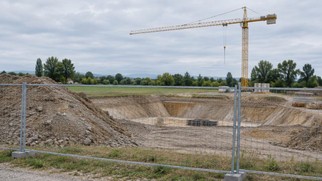 Symbolbild: eine eingezäunte Baustelle mit ausgehobener Baugrube und Kran am Ortsrand, dahinter Wiese und Baumreihe