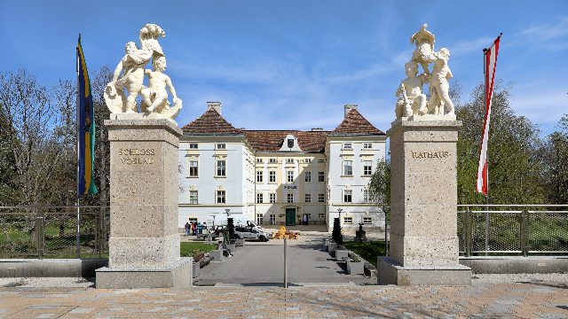 Zufahrt zum Schloss Vöslau mit zwei Steinpfeilern und Puttenfiguren, auf einem steht „Rathaus“, dahinter das weiße Schloss