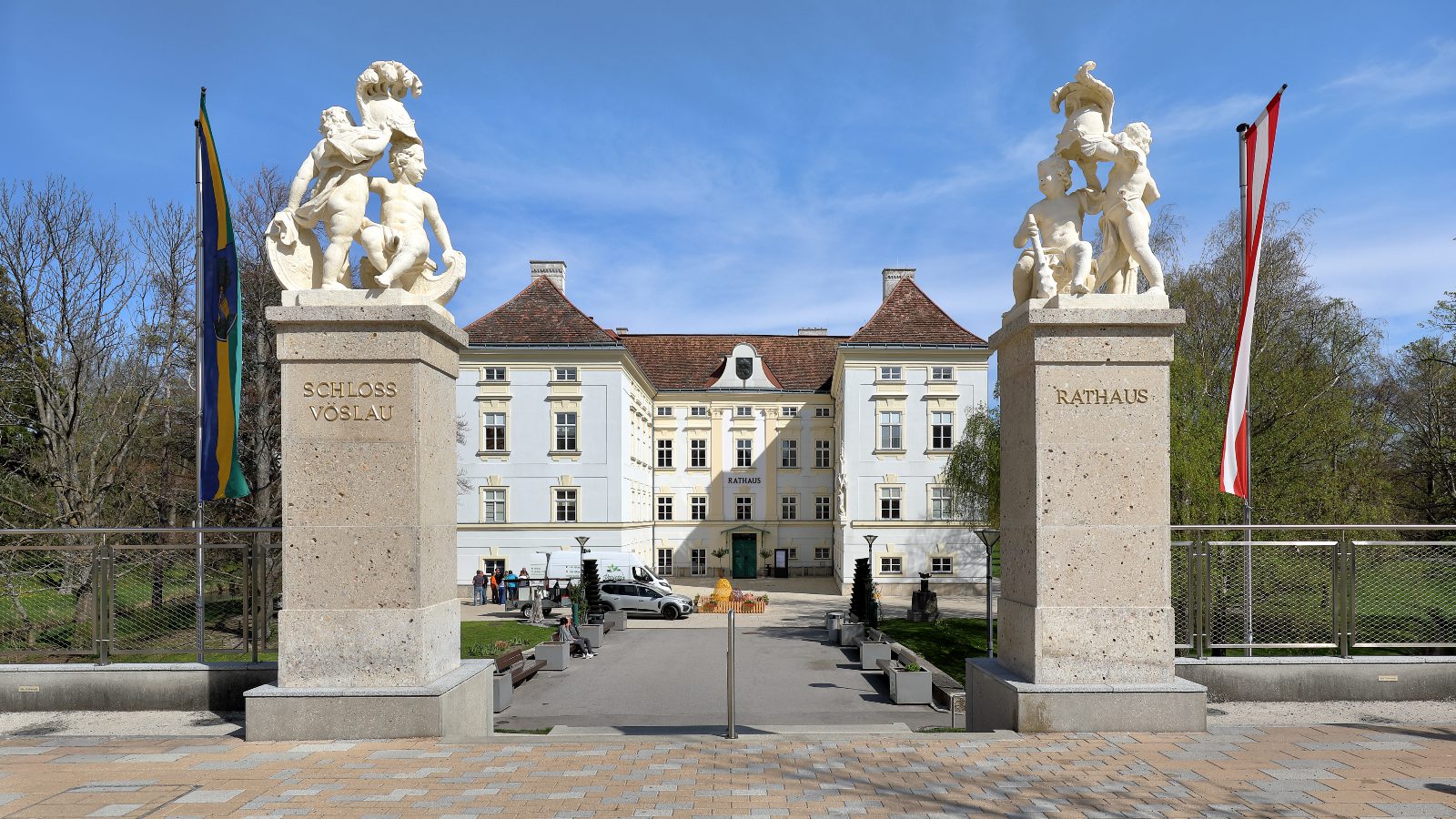 Zufahrt zum Schloss Vöslau mit zwei Steinpfeilern und Puttenfiguren, auf einem steht „Rathaus“, dahinter das weiße Schloss