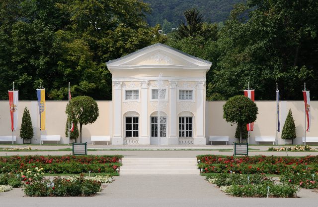 Weißer klassizistischer Pavillon vor einer hellen Parkmauer, davor Rosenbeete, Bänke und Fahnen, dahinter bewaldete Hügel