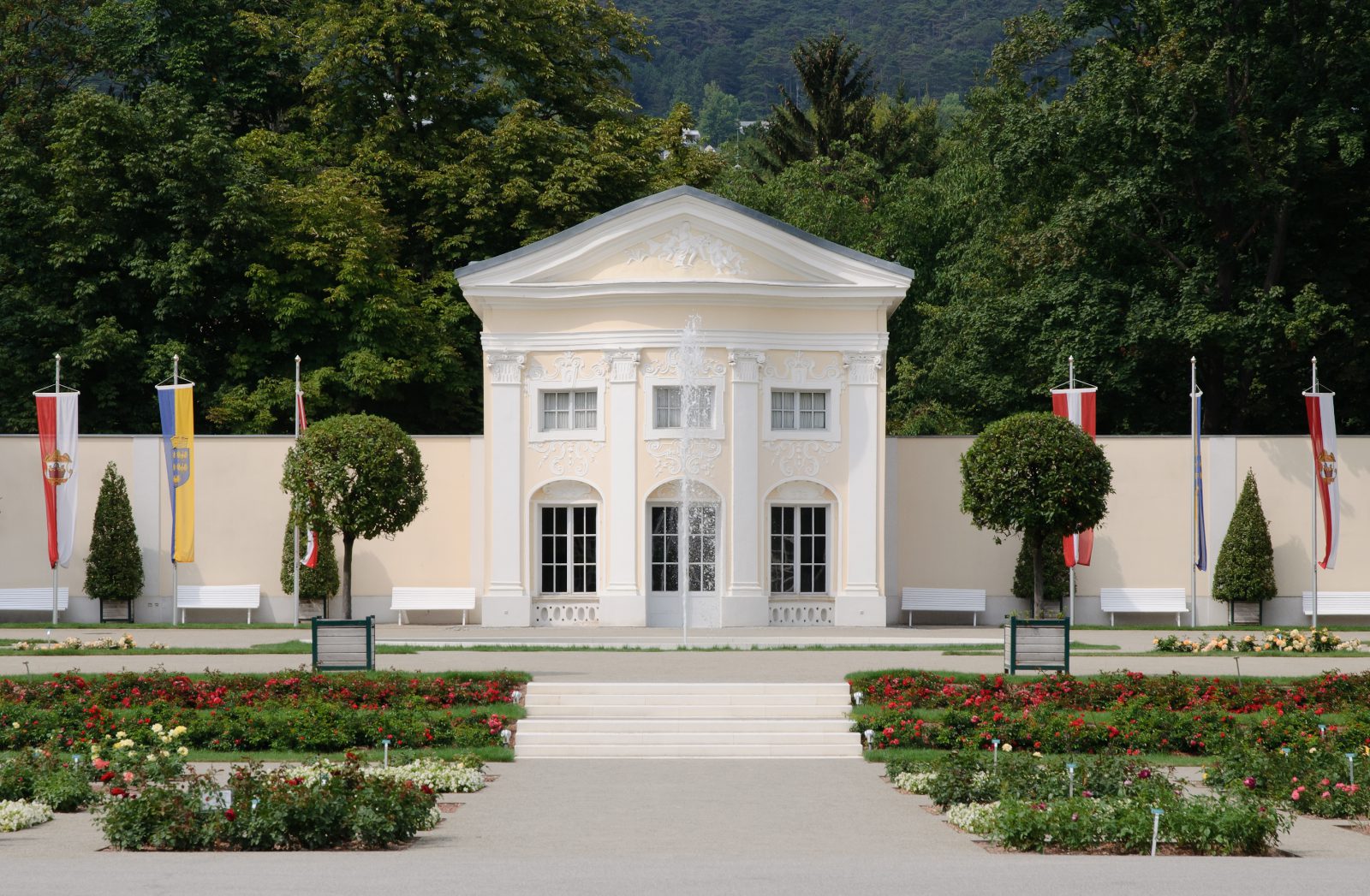 Weißer klassizistischer Pavillon vor einer hellen Parkmauer, davor Rosenbeete, Bänke und Fahnen, dahinter bewaldete Hügel