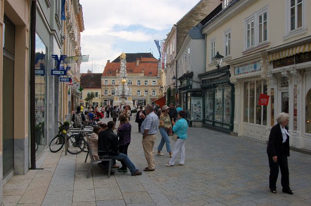 Fußgängerzone mit Geschäften und Passanten, am Ende die weiß-goldene Pestsäule vor einem Gebäude mit rotem Dach