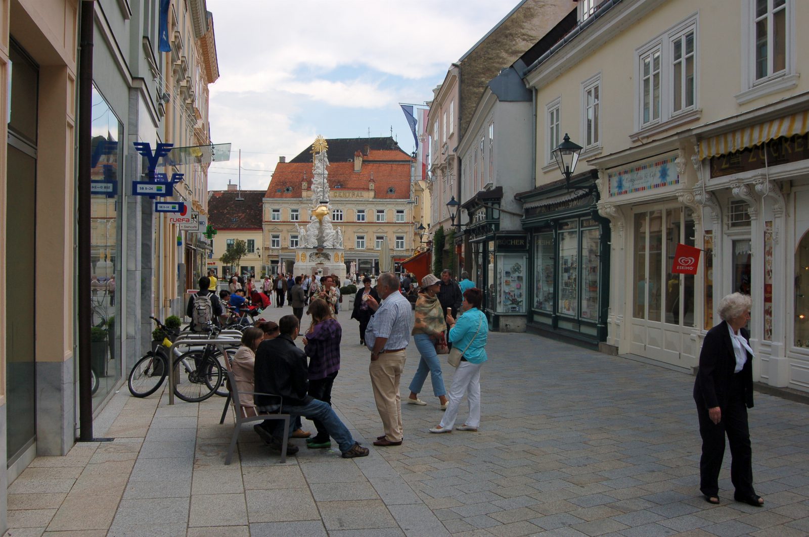 Fußgängerzone mit Geschäften und Passanten, am Ende die weiß-goldene Pestsäule vor einem Gebäude mit rotem Dach