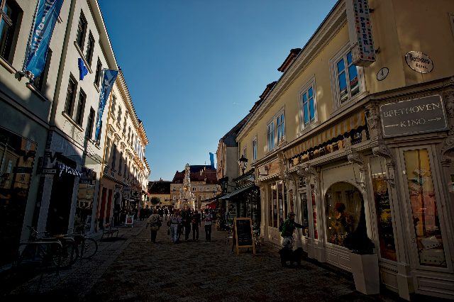 Blick durch die Fußgängerzone in Baden zur Dreifaltigkeitssäule am Hauptplatz, rechts das Schild zum Beethoven-Kino, Passanten unter blauem Himmel
